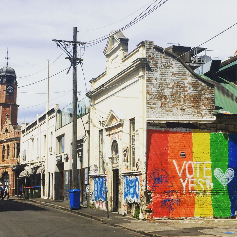 A laneway in Newtown. The side of the closest building is painted the colours of the rainbow, with the words "Vote Yes" written on top.