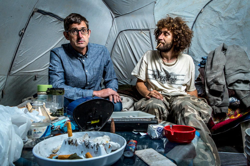 Theroux sits with Nate Walsh inside a tent. In the foreground you can see a full ashtray. The tent is messy and Walsh is unkempt.