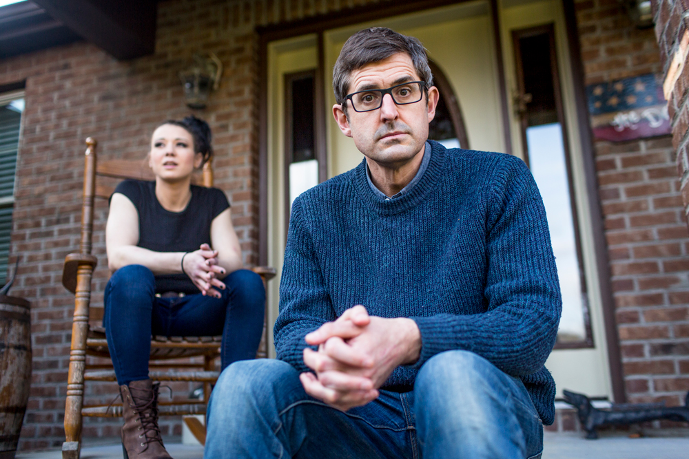 Theroux sits on the steps of a porch. Behind him, a young woman (Katillia) sits on a chair.