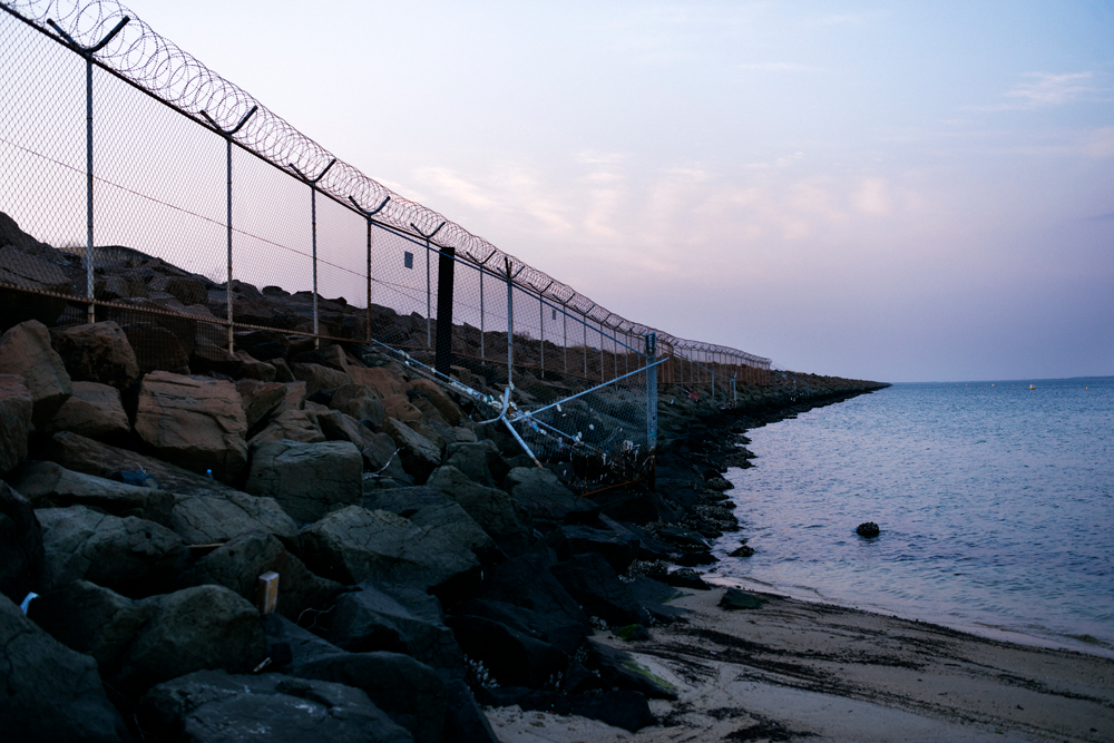 A fence with curled barb wire on top stretches along the rocks and into the distance, blocking access to Sydney Airport from the beach.